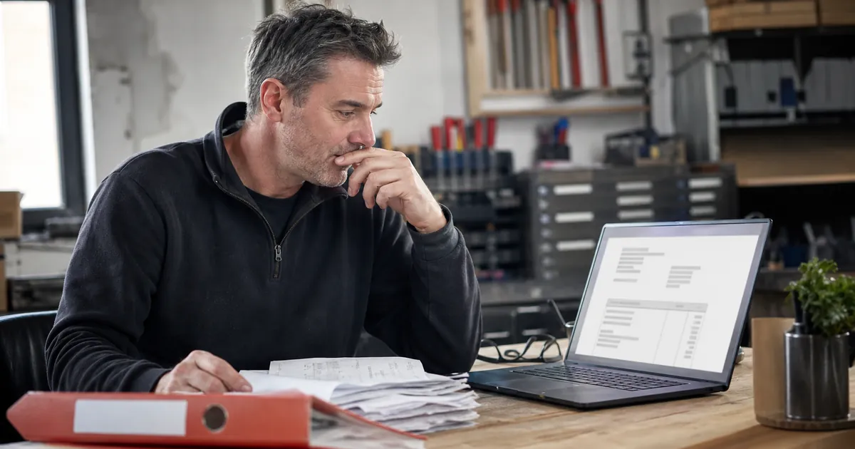 Handwerker prüft Rechnungen und Belege am Laptop in seiner Werkstatt