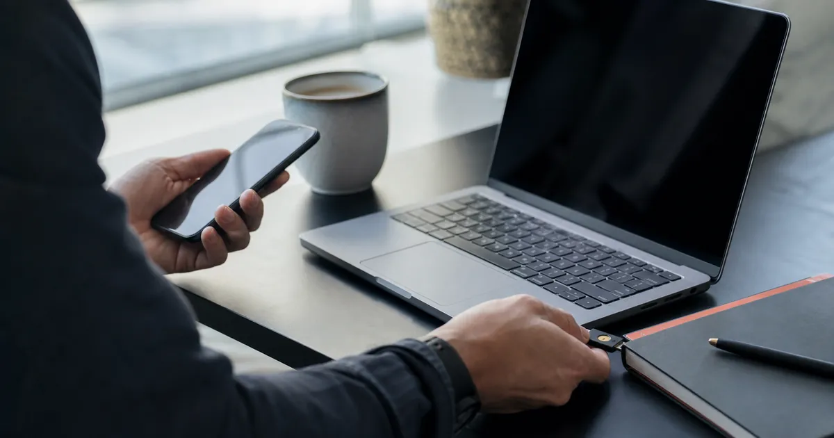 Hand hält Smartphone neben einem Laptop mit gesperrtem, dunklem Bildschirm als Sinnbild für einen abgesicherten Login